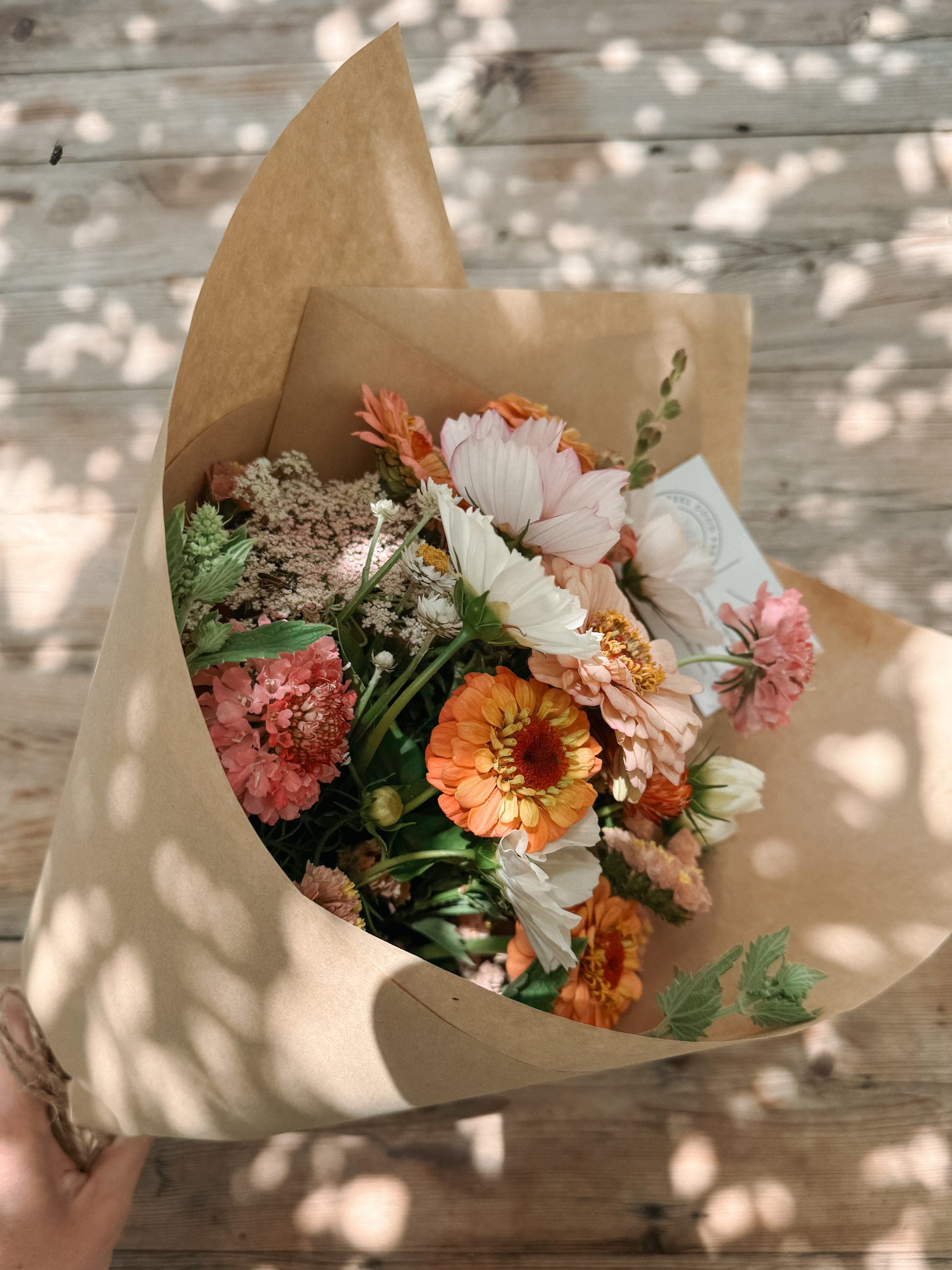 A farm bouquet wrapped in kraft paper, resting on weathered timber in dappled light.