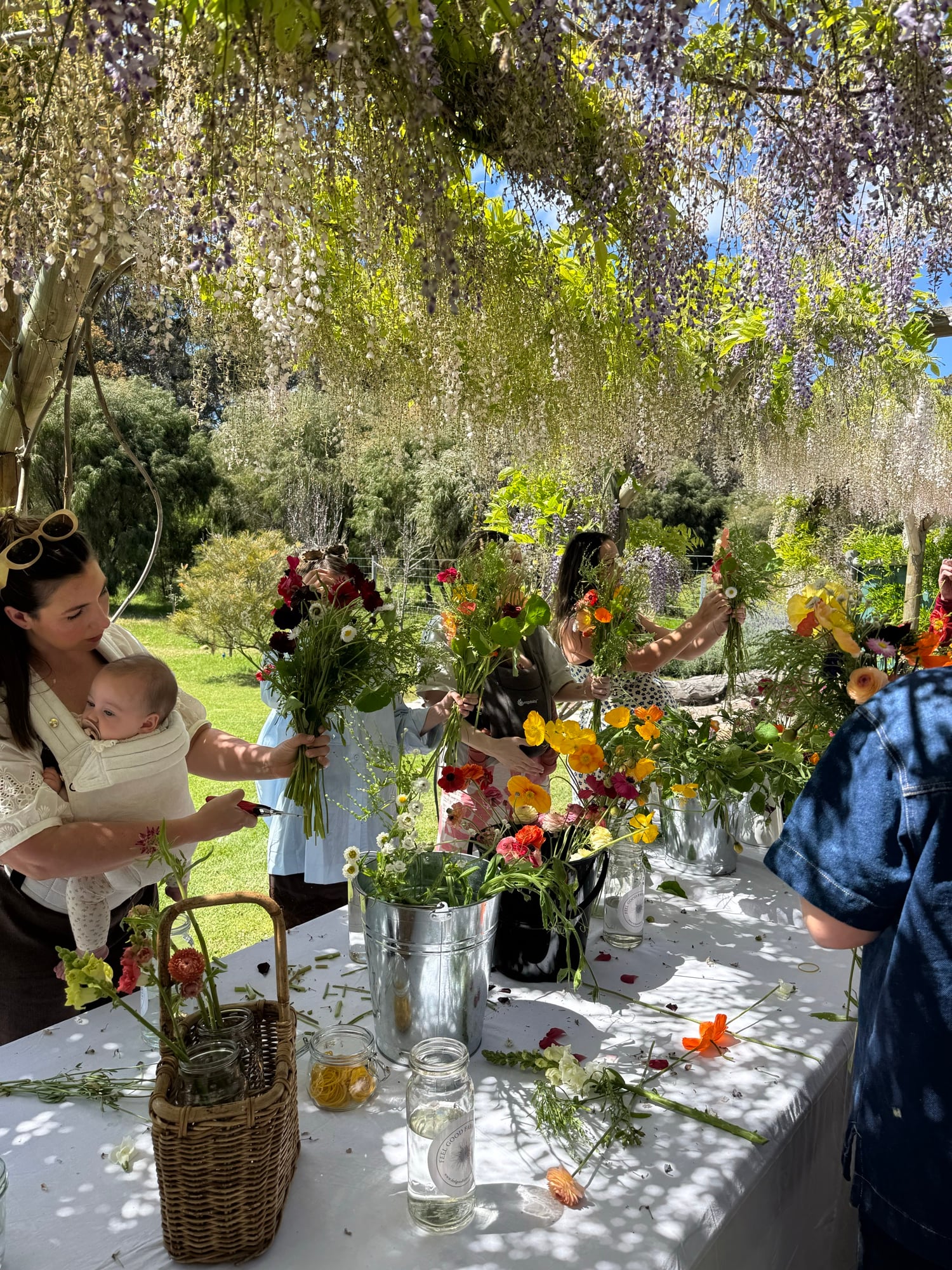 A long table under the wisteria at Feel Good Farm, guests holding up the bouquets they’ve made, buckets of poppies and ranunculus between them.