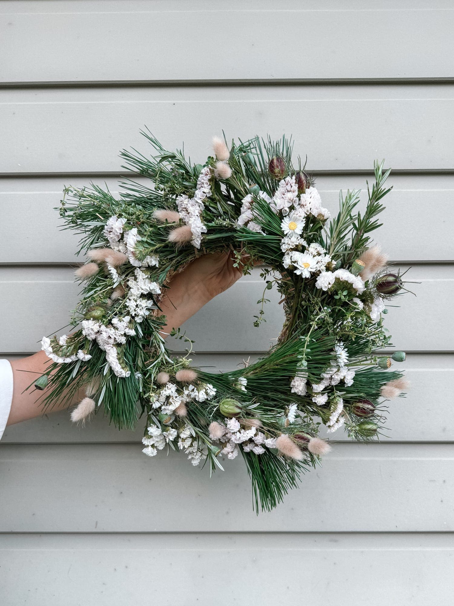 A hand holding up a just-made Christmas wreath — pine, rosemary, white statice, daisies and bunny tails.
