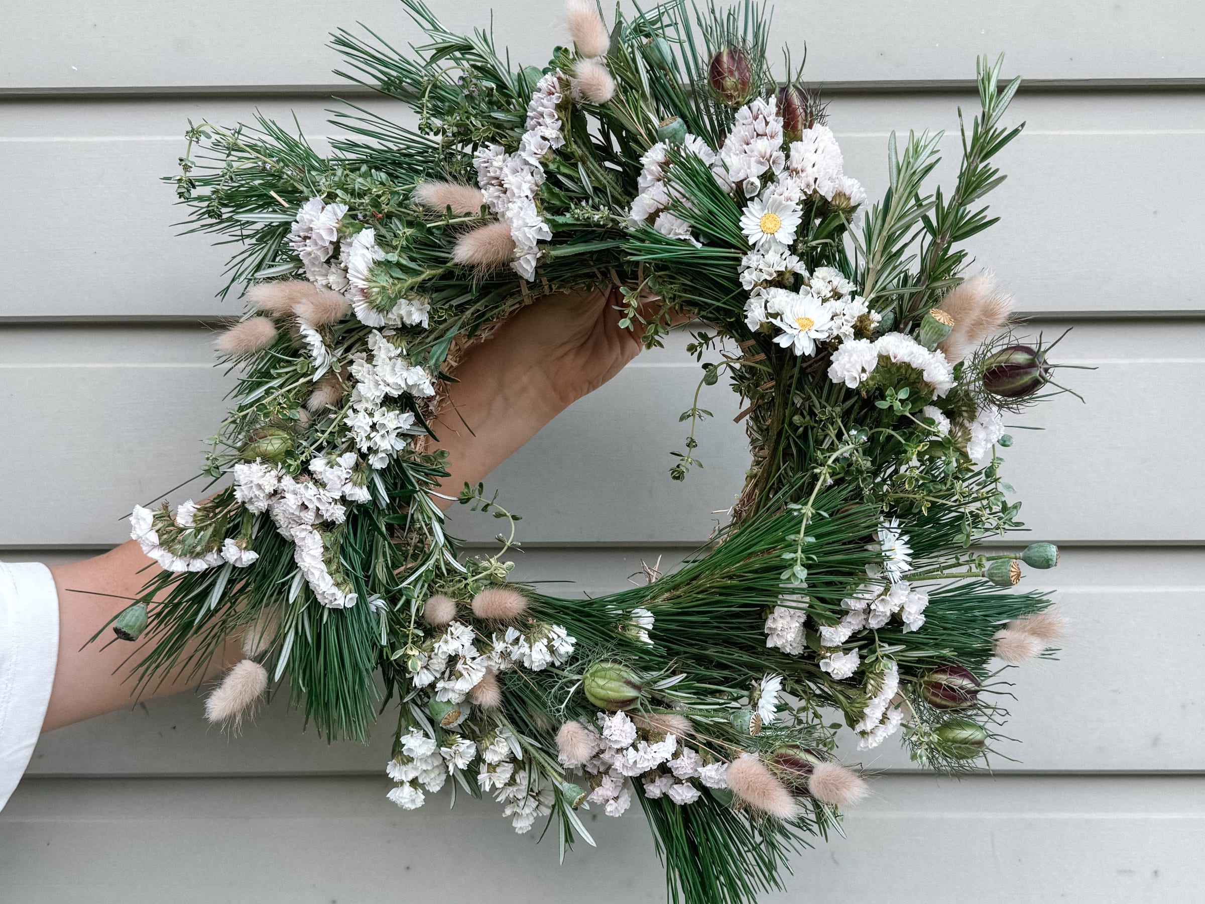 A Christmas wreath of pine, rosemary, white statice, daisies and bunny tails, held up against a pale weatherboard wall.